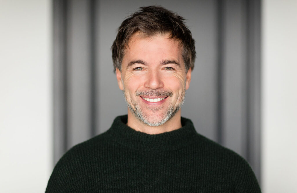 Smiling middle-aged man with a defined jawline and short salt-and-pepper stubble in a dark green sweater, representing men considering a deep plane facelift in Newport Beach