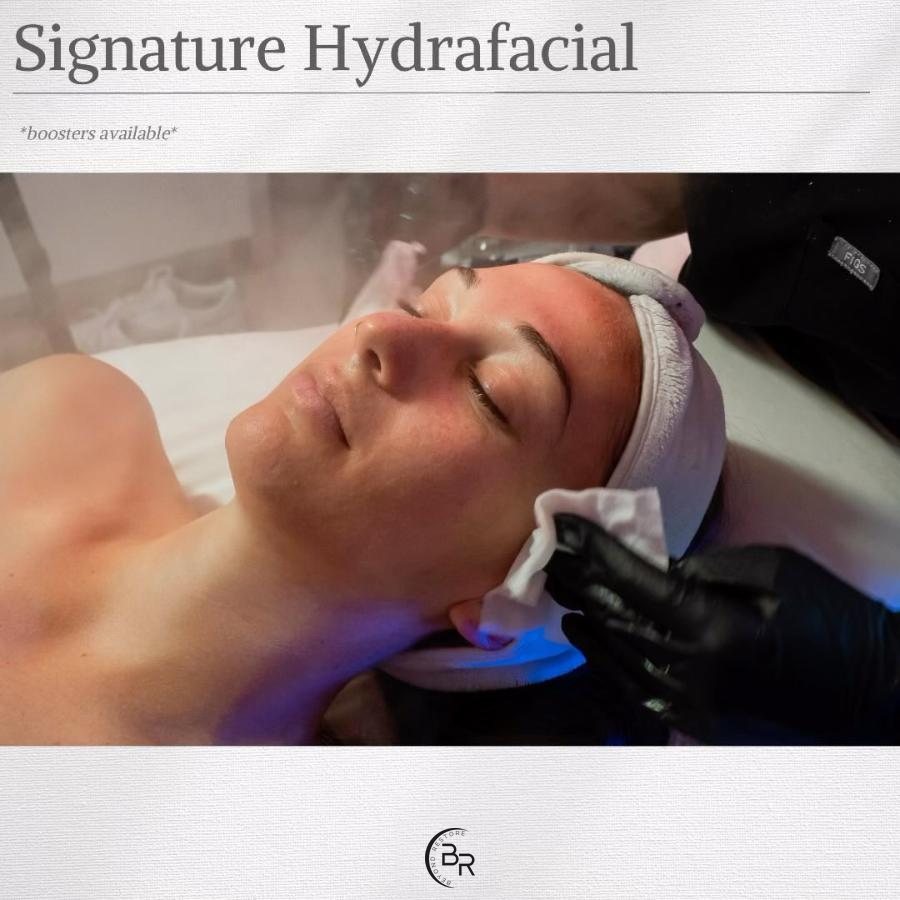 Woman relaxing with her eyes closed and her hair in a headband while a gloved aesthetician dabs her temple with a soft white cloth, with the words Signature HydraFacial above and the Beyond Restore logo below
