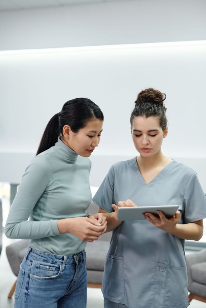 Woman in scrubs showing a tablet screen to a woman in a gray turtleneck in a bright room