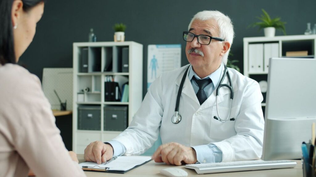 Doctor in a white coat with a stethoscope talking with a woman across a desk in a bright office