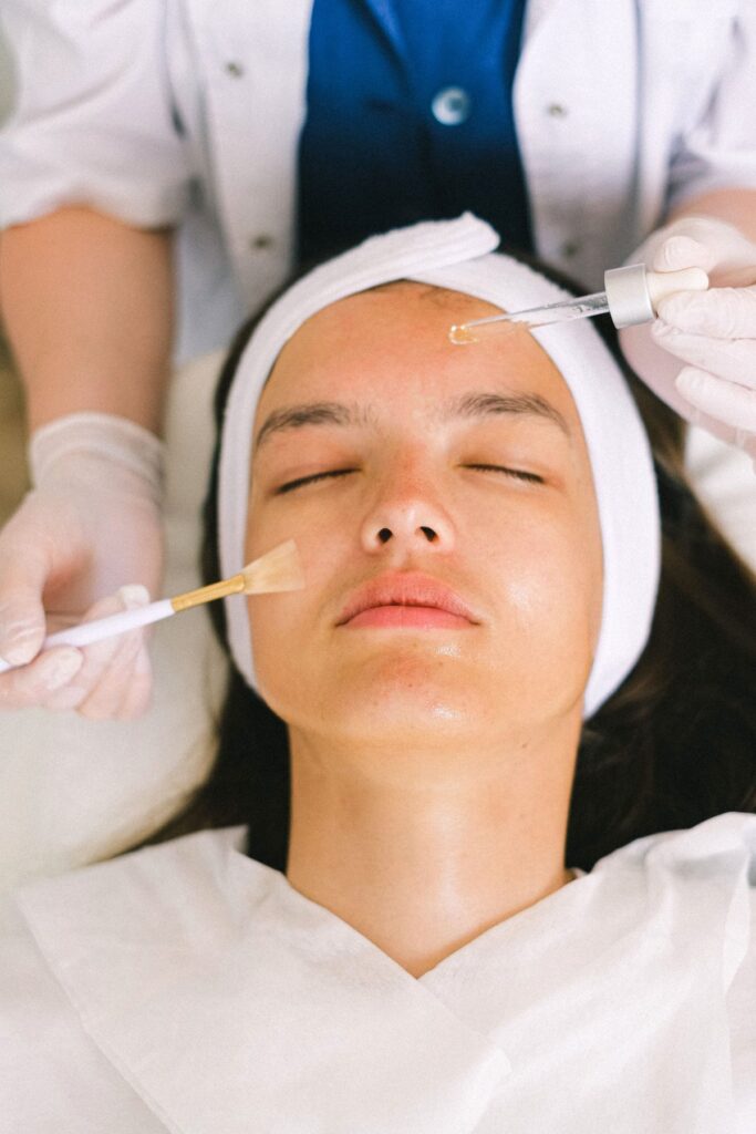 A provider applying a cosmetic product to a client's face with a brush while the client rests with a headband on, illustrating a facial treatment visit