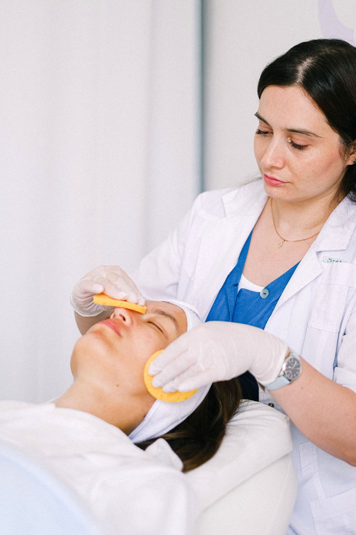 Skincare professional in a white coat gently cleansing a client's face with sponges during a facial
