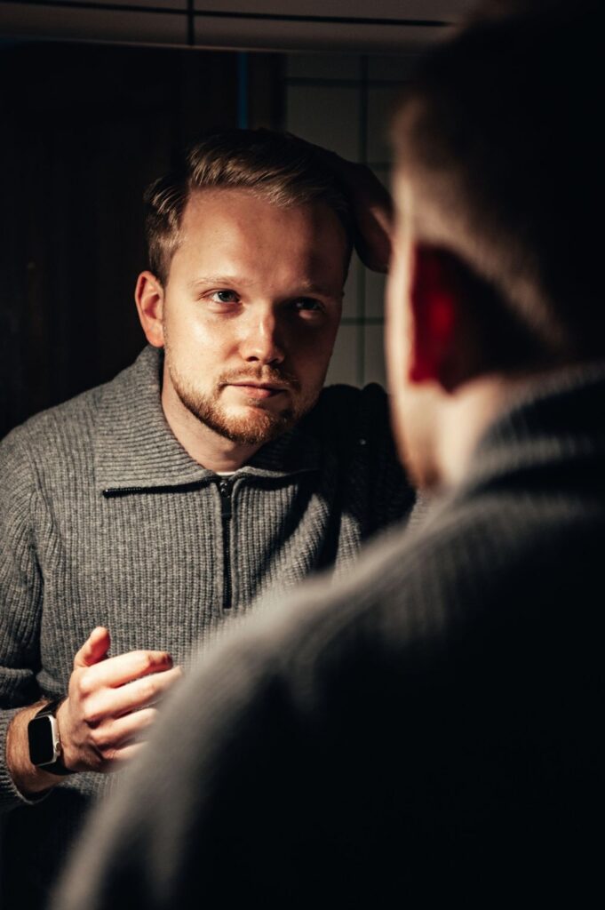 Man in a gray zip-neck sweater touching his hair while looking at his reflection in a mirror