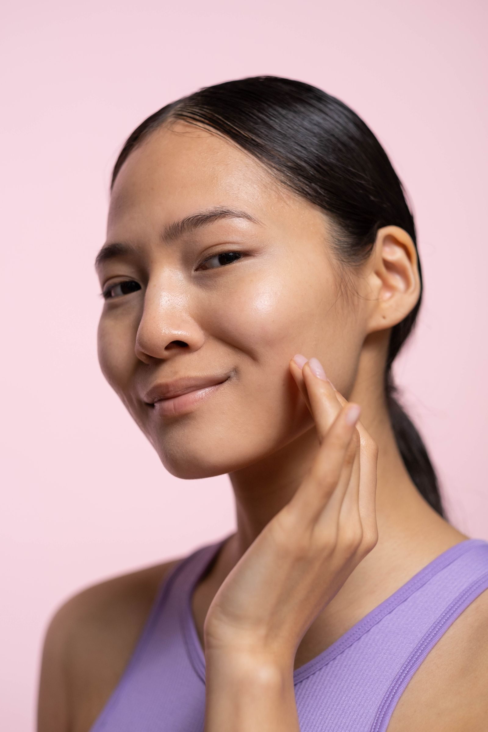 Asian woman with dewy, glowing skin touching her jawline against a soft pink background, illustrating smooth skin goals with Botox
