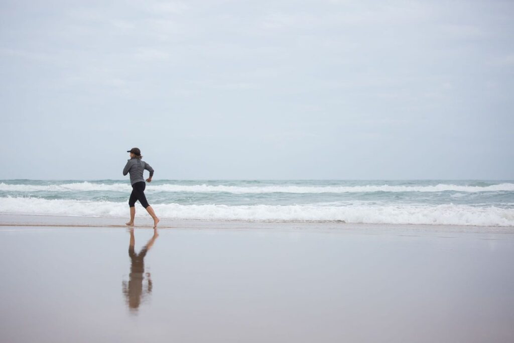 Woman jogging barefoot along the shoreline on an overcast day, her reflection on the wet sand
