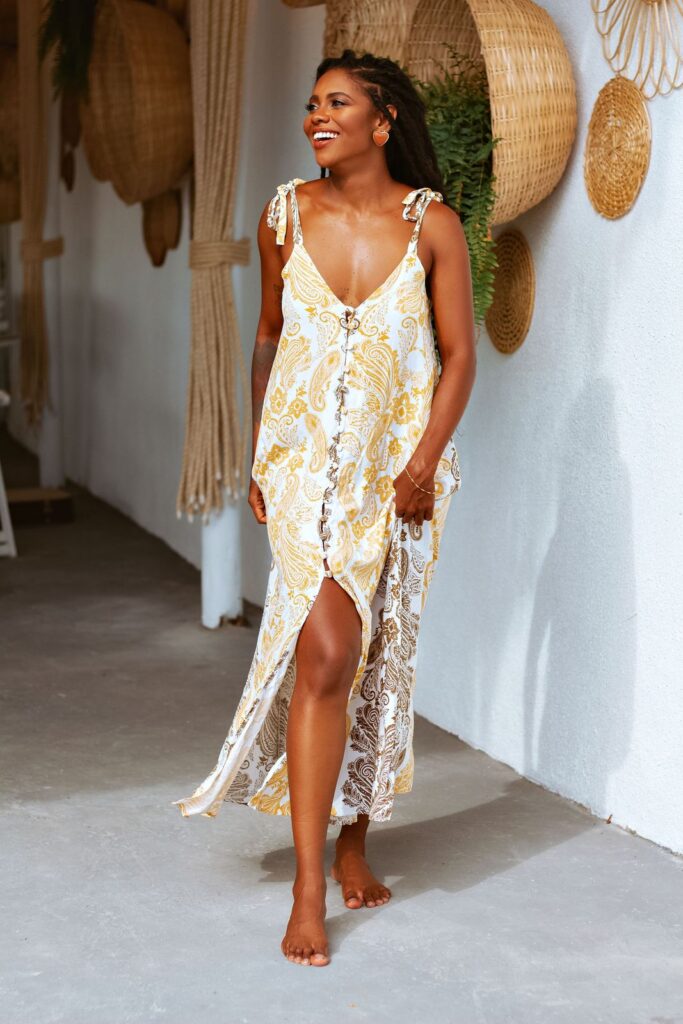 Smiling woman in a yellow and white patterned summer dress walking barefoot past a white wall with woven baskets