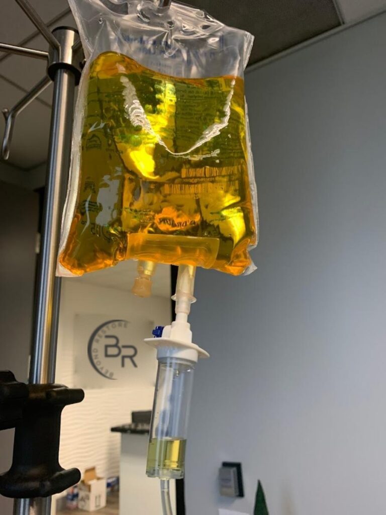 Yellow IV vitamin bag hanging from an IV pole in the Beyond Restore office, with the Beyond Restore logo on the wall behind it