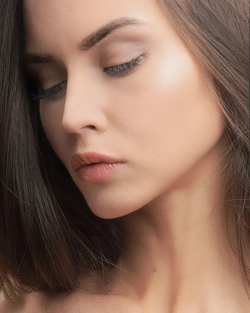 Close-up of a woman with lowered eyes and a smooth, defined jawline against a soft neutral background