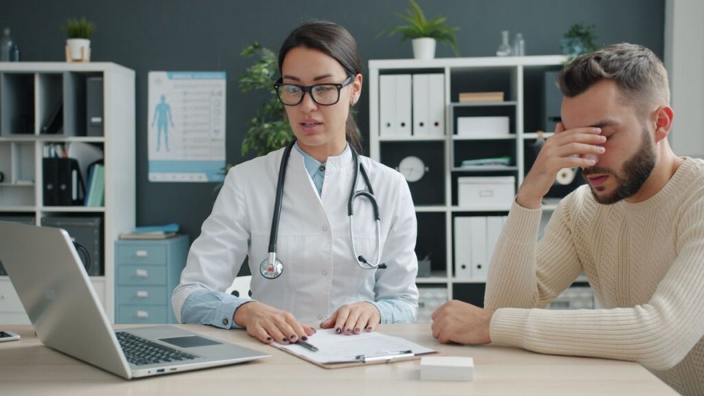 Doctor in a white coat taking notes while talking with a man across a desk in a bright office