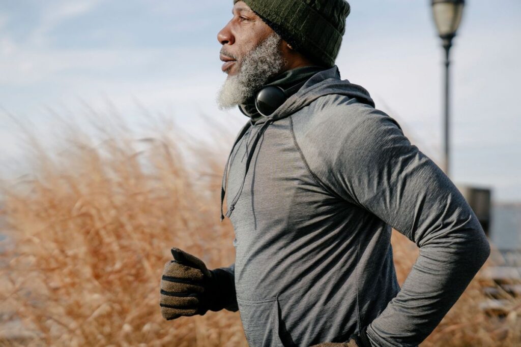 Bearded man in a gray hoodie and beanie jogging outdoors past tall dry grass